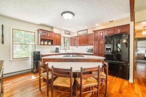 A kitchen with wooden cabinets, a black refrigerator, a large island with chairs, and a window with a view of trees.