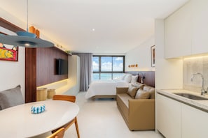 Una habitación de hotel moderna con cama, sofá, mesa de comedor, zona de cocina y un amplio ventanal con vistas.