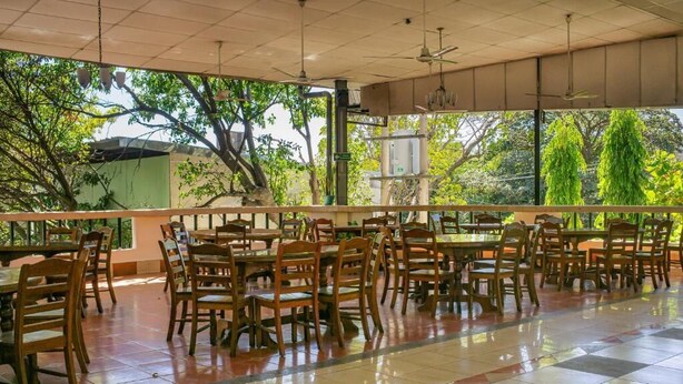 A spacious indoor dining area with wooden tables and chairs, large windows with a view of trees, and ceiling fans.