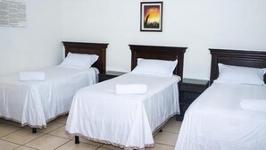 A hotel room with three single beds, each with a white bedspread and a pillow. There is a framed picture on the wall above the beds.
