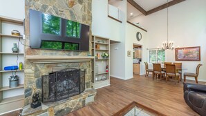 A living room with a stone fireplace, a flat-screen TV, wooden flooring, a dining area with chairs, and a chandelier. - Cuesta Verde at Rumbling Bald (Lake Lure)