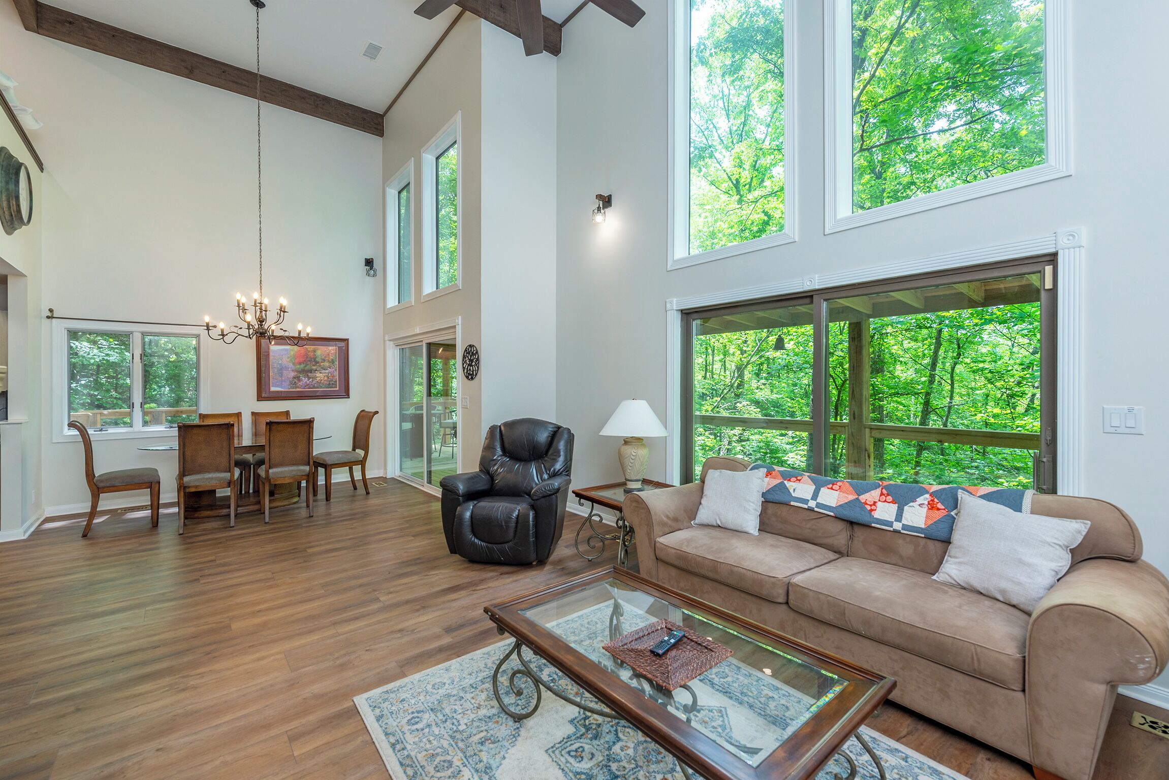 A spacious living room with a wooden floor, a leather chair, a beige sofa, a glass coffee table, and large windows offering a view of greenery.
