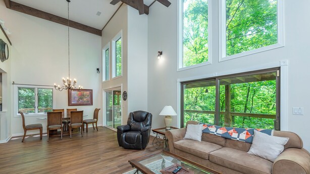 A spacious living room with a wooden floor, a leather chair, a beige sofa, a glass coffee table, and large windows offering a view of greenery. - Cuesta Verde at Rumbling Bald (Lake Lure)