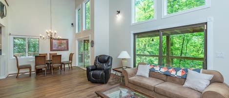 A spacious living room with a wooden floor, a leather chair, a beige sofa, a glass coffee table, and large windows offering a view of greenery.
