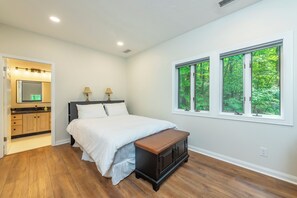 A bedroom with a bed, bedside lamps, a wooden bench, a vanity with a mirror, and a window with a view of greenery. - Cuesta Verde at Rumbling Bald (Lake Lure)