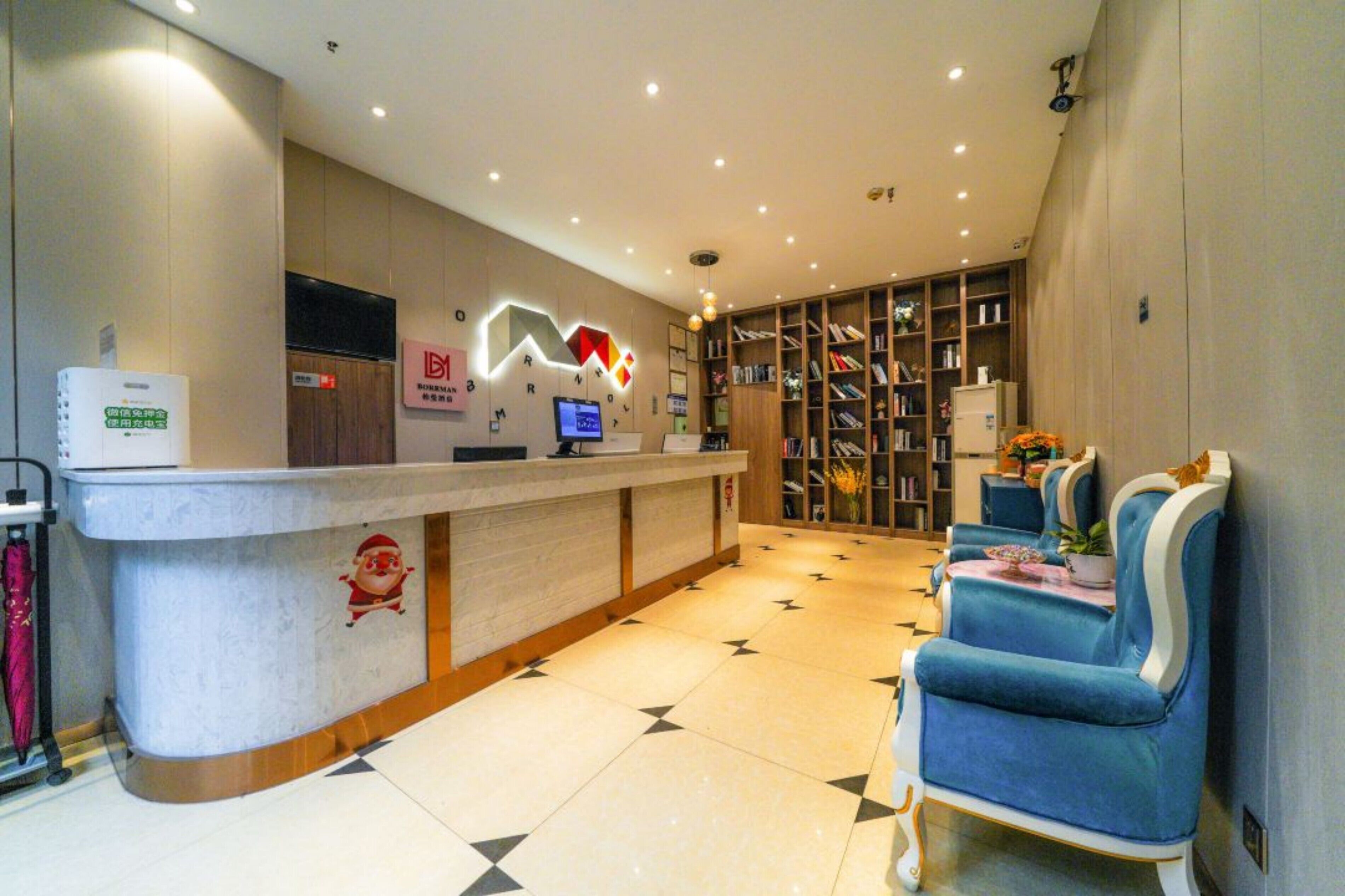 Zona de recepción de hotel con mostrador, computadora, estantería y sillas azules.