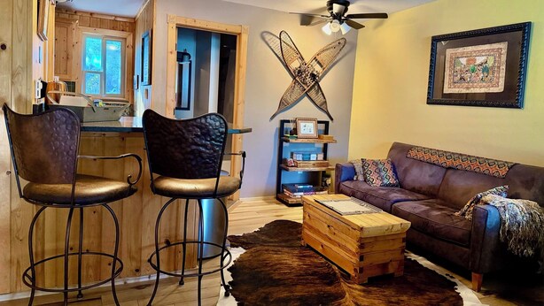 Ein gemütliches Wohnzimmer mit einer Couch, zwei Barhockern, einem Couchtisch, einem Kuhfellteppich, einem Deckenventilator und einer sichtbaren Küche im Hintergrund.