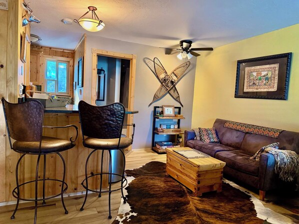 A cozy living room with a sofa, two bar stools, a coffee table, a cowhide rug, a ceiling fan, and a kitchen visible in the background.