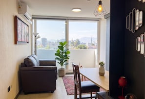 Una sala de estar moderna con sofá, mesa de comedor, sillas, una planta en maceta y vistas amplias a través de ventanas grandes.
