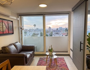 Ein modernes Wohnzimmer mit einer Ledersofa, einem Glastisch mit einem dekorativen Gegenstand, einem farbenfrohen Teppich und einer großen Schiebetür, die auf einen Balkon mit Blick auf die Stadt führt.