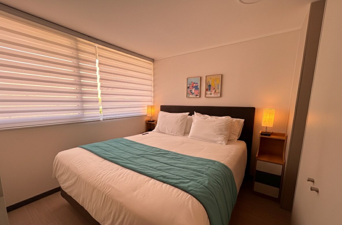 A hotel room with a bed, two bedside lamps, a window with blinds, and two wall-mounted pictures.