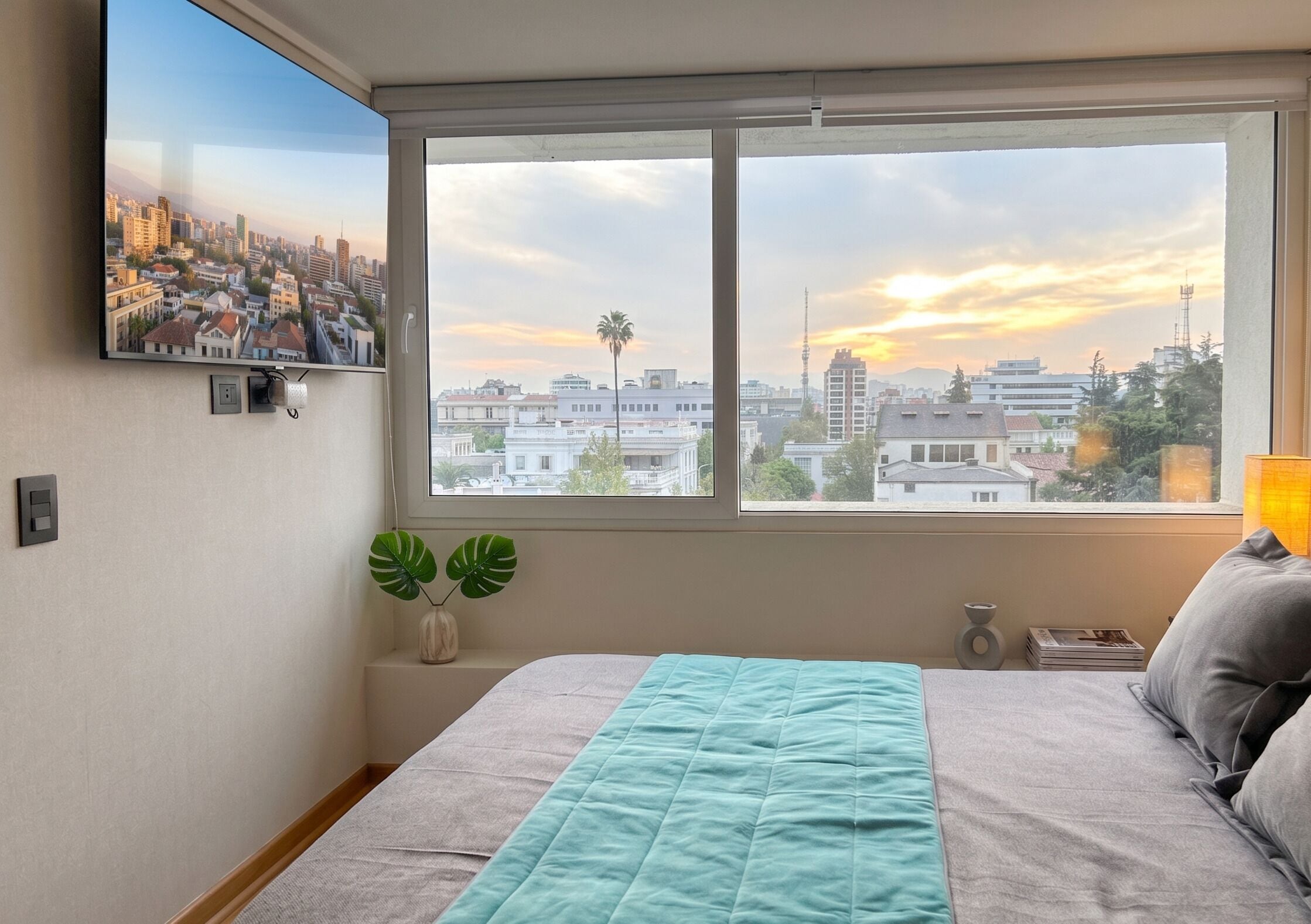 Un dormitorio con cama, ventana con vista panorámica de la ciudad, televisor montado en la pared y una planta.