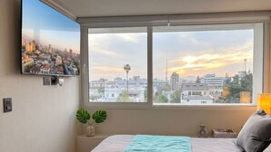 A bedroom with a bed, a window with a cityscape view, a wall-mounted TV, and a plant. - Centro Cívico - La Moneda - Domus Travels (Santiago)