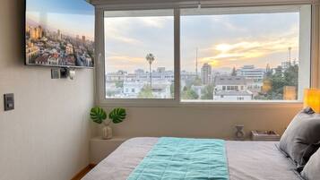 Un dormitorio con cama, ventana con vista panorámica de la ciudad, televisor montado en la pared y una planta.