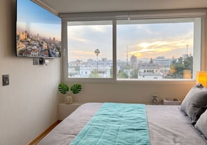 Un dormitorio con cama, ventana con vista panorámica de la ciudad, televisor montado en la pared y una planta.