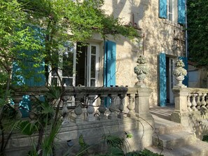 Eine Steinschutzmauer mit einem Brunnen, ein Gebäude mit blauen Fensterläden und ein Baum im Vordergrund.