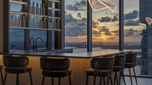 A modern bar area with a city skyline view at sunset, a large window, and a shelf with bottles. - Presidential Penthouse Private Infinity Pool (Miami)