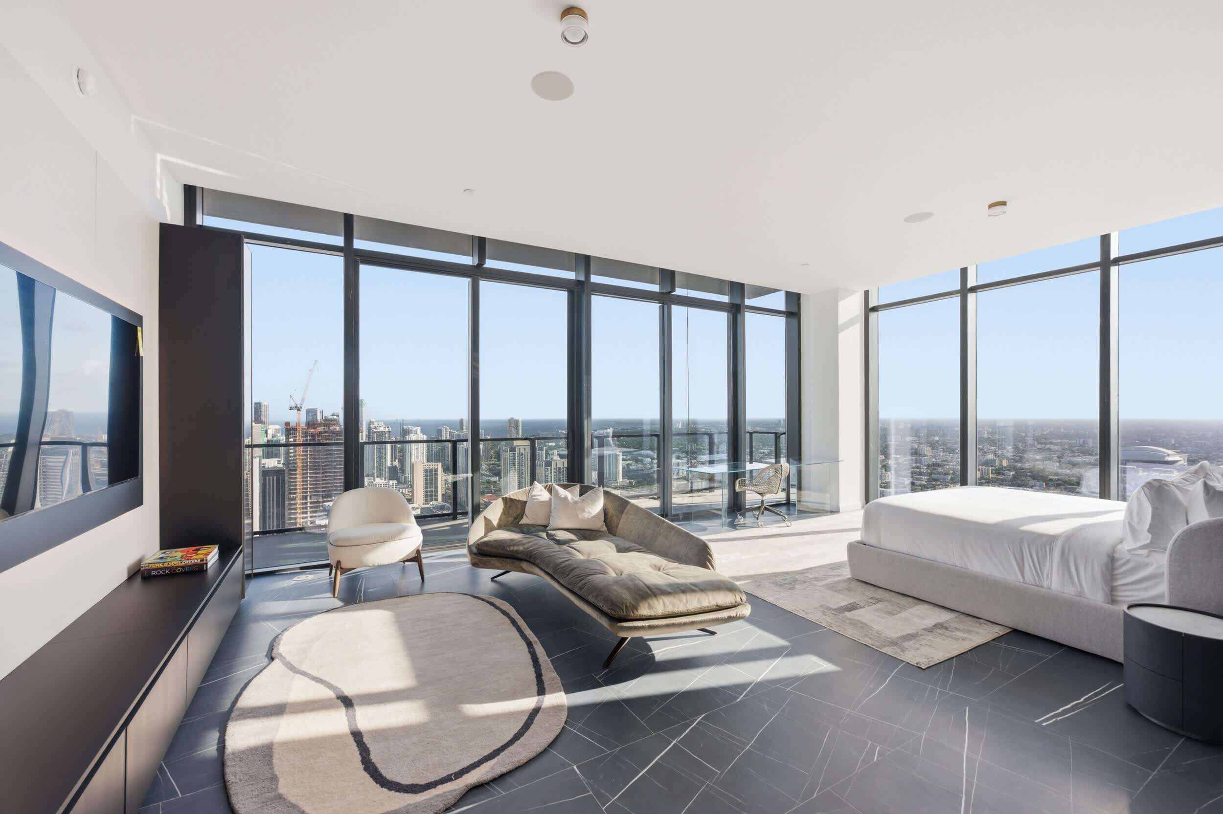 A modern bedroom with a large bed, a sofa, and a chair, with a city view through large windows.