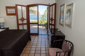 Ein Zimmer mit einem Bett, einem Sessel, einem Schreibtisch, einem Fenster mit Meerblick und einer Tür zu einem Außenbereich.