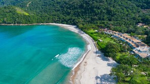 Luftaufnahme einer Küstenlandschaft mit Strand, klarem türkisblauem Wasser und einer Reihung von Häusern vor einer hügeligen Kulisse.