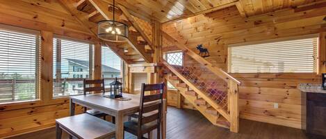 L’intérieur d’un chalet en bois, avec une table à manger, des chaises, un escalier et une vue sur l’extérieur.