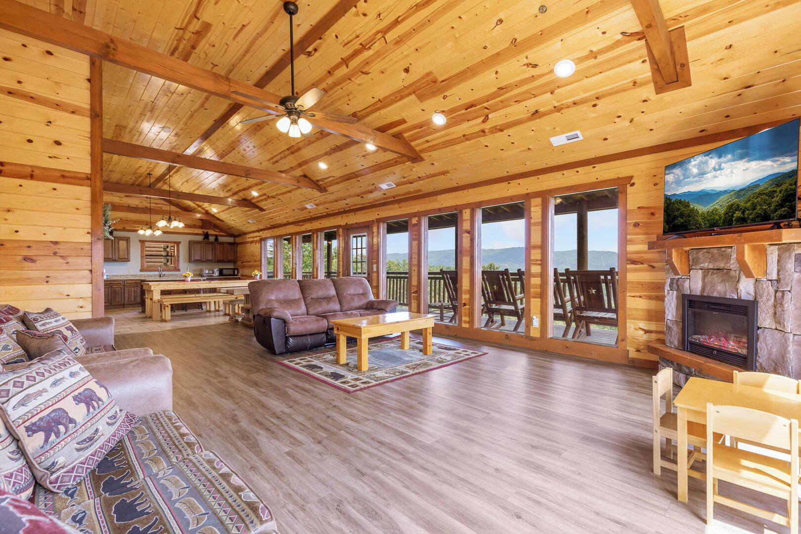 A log cabin interior with a fireplace, a sofa, a dining table, and a large window overlooking a mountainous landscape.