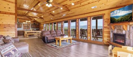 A log cabin interior with a fireplace, a sofa, a dining table, and a large window overlooking a mountainous landscape.