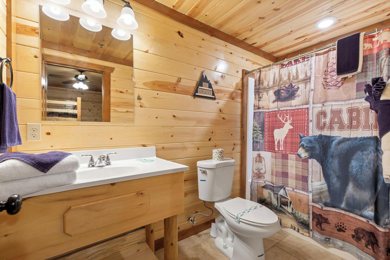 Une salle de bain rustique, aménagée dans une cabane en bois, avec un lavabo, des toilettes et un rideau de douche orné de motifs animaliers.