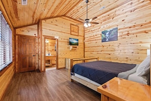 A wooden cabin interior with a bed, a ceiling fan, a TV, and a bathroom with a window. - A Luxury Mountain Oasis for Families, Retreats & Group Getaways (Sevierville)