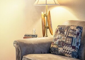 Ein gemütlicher Winkel mit einer Lampe, einem Kissen mit Patchwork-Muster und einem Stapel Bücher auf einem Sessel.