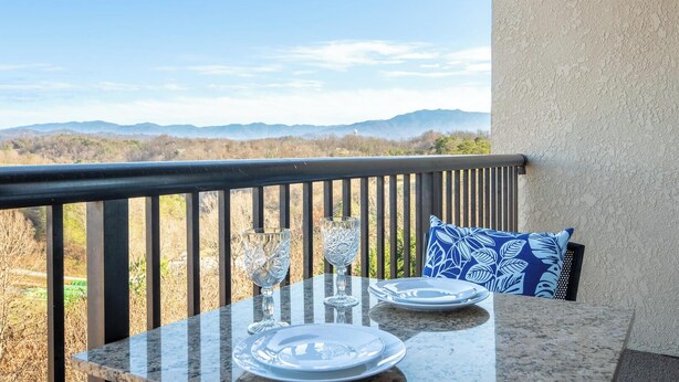 Un balcón con una mesa preparada para dos personas, un cojín estampado en azul y vistas a montañas distantes.