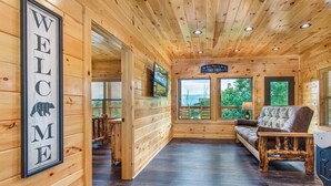 Een houten cabin interieur met een 'WELCOME' bord, een bank, een televisie en een raam met uitzicht op groen.