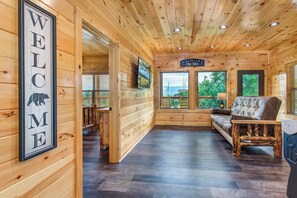 Innenansicht einer Holzhütte mit einem 'WELCOME'-Schild, einer Couch, einem Fernseher und einem Fenster mit Blick ins Grüne.