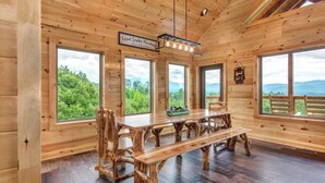 Interieur van een houten hutje met een grote eettafel, meerdere stoelen en grote ramen met uitzicht op een bebost landschap.