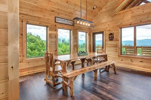 Ein Holzblockhaus-Interieur mit einem großen Esstisch, mehreren Stühlen und großen Fenstern, die Blick auf eine bewaldete Landschaft bieten.