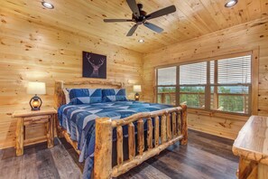 A wooden cabin bedroom with a bed, two bedside lamps, a ceiling fan, a window with blinds, and a deer antler artwork on the wall. - Jump On Inn: Luxury Smoky Mountain Cabin With Indoor Pool (Sevierville)