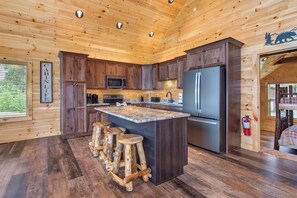 Ein Interieur einer Hütte mit einer Küche, die Holzschränke, eine zentrale Kochinsel mit Hockern, einen Edelstahlkühlschrank und einen fest montierten Feuerlöscher zeigt.