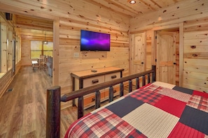 Interior de una cabaña de troncos con cama, televisor montado en la pared y zona de comedor con muebles de madera.