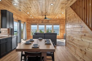 Ein Holzinterieur mit einem Esstisch für vier Personen, einer Couch und Blick auf Berge durch große Fenster.