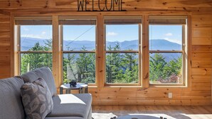 Interieur van een houten hutje met een 'WELCOME'-bord, een grijze bank en grote ramen met uitzicht op bergen.