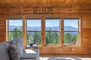 Ein Holzhütten-Innenraum mit einem 'WELCOME'-Schild, einem grauen Sofa und großen Fenstern mit Blick auf die Berge.
