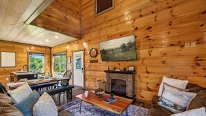 A log cabin interior with a fireplace, wooden walls, a dining area, and a large window. - New! Indoor Pool, Game Room, Families Welcome! (Sevierville)