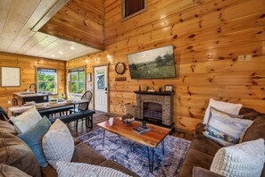 A log cabin interior with a fireplace, wooden walls, a dining area, and a large window. - New! Indoor Pool, Game Room, Families Welcome! (Sevierville)