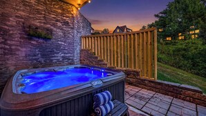 A hot tub with a stone wall backdrop, wooden deck, and a view of a residential area at dusk. - New! Indoor Pool, Game Room, Families Welcome! (Sevierville)