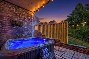 A hot tub with a stone wall backdrop, wooden deck, and a view of a residential area at dusk. - New! Indoor Pool, Game Room, Families Welcome! (Sevierville)