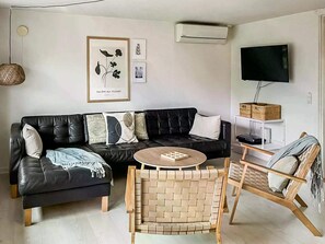 Ein modernes Wohnzimmer mit einem schwarzen Ledersofa, einem runden Couchtisch aus Holz, einem gewebten Sessel, einem Flachbildfernseher und einer Wand-Klimaanlage.