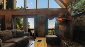 A cozy cabin interior with a stone fireplace, a wooden table, a leather sofa, and a large window offering a view of the outdoors. - A-Frame Mountain Retreat | Jaw-Dropping Smoky Mountain Views, Hot Tub & Arcade (Pigeon Forge)