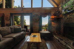 L’intérieur chaleureux d’un chalet avec une cheminée en pierre, une table en bois, un canapé en cuir et une grande fenêtre donnant sur le paysage extérieur.
