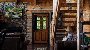 A log cabin interior with a stone fireplace, wooden walls, a wooden door, leather furniture, and a view of the outdoors. - A-Frame Mountain Retreat | Jaw-Dropping Smoky Mountain Views, Hot Tub & Arcade (Pigeon Forge)