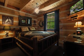 Ein Schlafzimmer in einer Blockhütte mit Bett, Holzwänden, einer Deckenleuchte, einem Fernseher und einem Fenster mit Blick auf Grünflächen.